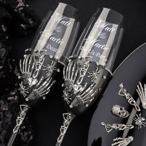 Op de afbeelding: Twee zwarte champagnefluiten versierd met zilveren skelethandjes, spinnen en strass. De fluiten zijn gegraveerd met de tekst "Matt & Martie Till Death" en "Matt & Martie Do Us Part".