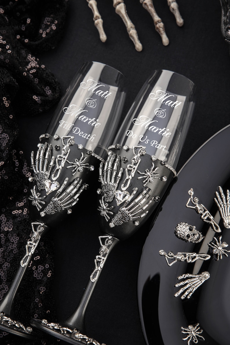Op de afbeelding: Twee zwarte champagnefluiten versierd met zilveren skelethandjes, spinnen en strass. De fluiten zijn gegraveerd met de tekst "Matt & Martie Till Death" en "Matt & Martie Do Us Part".