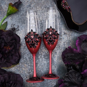May include: Two burgundy-colored champagne flutes with black and red crystal embellishments. The flutes are personalized with the names "Autumn" and "Jesse" and the date "11.23.2023".