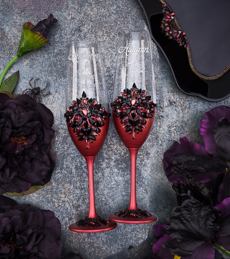May include: Two burgundy-colored champagne flutes with black and red crystal embellishments. The flutes are personalized with the names "Autumn" and "Jesse" and the date "11.23.2023".