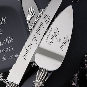 Op de afbeelding: Een zwarte en zilveren bruidstaartmes- en -serverset met een skelethandontwerp. Het mes heeft de tekst "Till death do us part" en de server heeft de tekst "Matt & Martie 10/13/2023".
