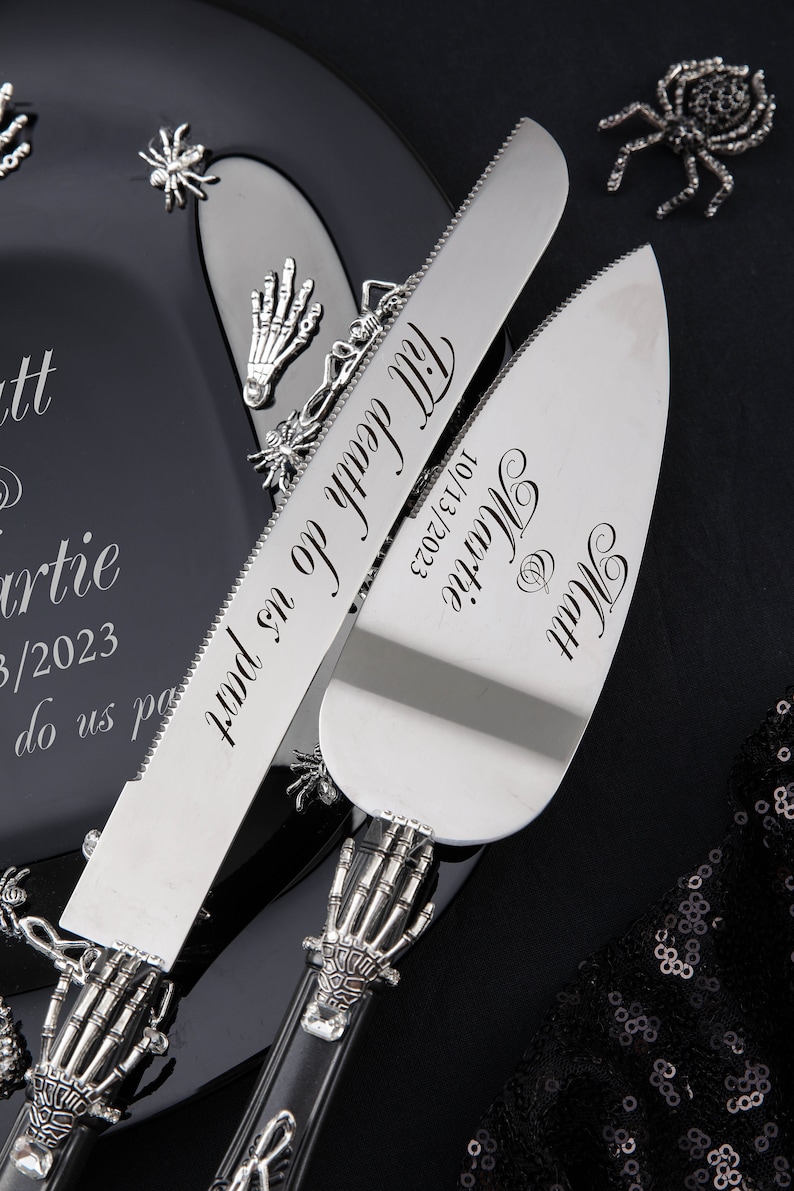 Op de afbeelding: Een zwarte en zilveren bruidstaartmes- en -serverset met een skelethandontwerp. Het mes heeft de tekst "Till death do us part" en de server heeft de tekst "Matt & Martie 10/13/2023".