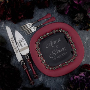May include: A red cake plate with a black glass insert featuring the names Anna & Steven and the date 09/08/2023. The plate is surrounded by a decorative border of silver leaves and rhinestones. Two silver cake knives with black handles are included. One knife is engraved with "Me & Mrs. Thomas 09/05/2023" and the other is engraved with "Til Death Do Us Part."