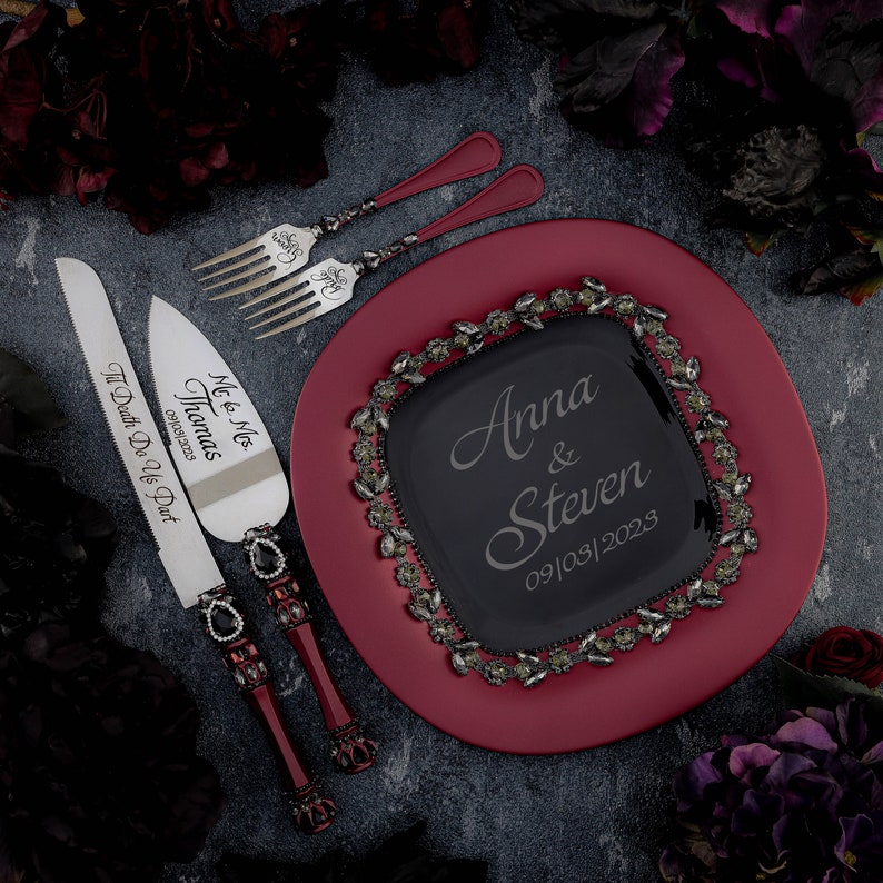 May include: A red cake plate with a black glass insert featuring the names Anna & Steven and the date 09/08/2023. The plate is surrounded by a decorative border of silver leaves and rhinestones. Two silver cake knives with black handles are included. One knife is engraved with "Me & Mrs. Thomas 09/05/2023" and the other is engraved with "Til Death Do Us Part."
