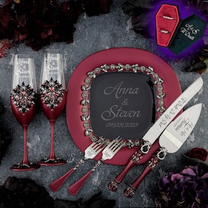 May include: A burgundy cake plate with a black square plate in the center, engraved with the names "Anna & Steven" and the date "09/03/2023". Two champagne flutes with rhinestones and the names "Anna & Steven" engraved on them. Two cake knives with black handles and rhinestones, engraved with "Mr. & Mrs. Thomas" and "Til Death Do Us Part". A black coffin-shaped ring box with a green velvet interior.
