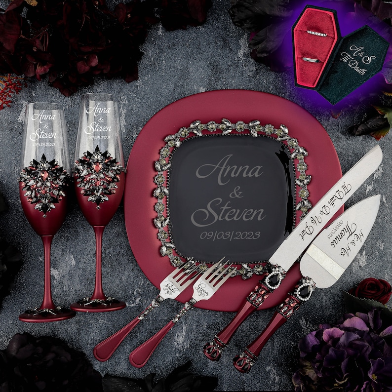 May include: A burgundy cake plate with a black square plate in the center, engraved with the names "Anna & Steven" and the date "09/03/2023". Two champagne flutes with rhinestones and the names "Anna & Steven" engraved on them. Two cake knives with black handles and rhinestones, engraved with "Mr. & Mrs. Thomas" and "Til Death Do Us Part". A black coffin-shaped ring box with a green velvet interior.
