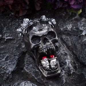 May include: A black skull ring holder with a silver crown of flowers and the words "A & H Till Death" on the forehead. Two silver skull rings with red gems are inside the skull's mouth.