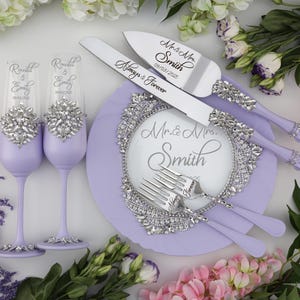 May include: A lavender wedding cake serving set with a knife, two forks, and two champagne flutes. The knife and forks have silver handles with rhinestones and the flutes have silver bands with rhinestones. The cake plate is lavender with a silver rhinestone border and the text "Mr. & Mrs. Smith" in the center.