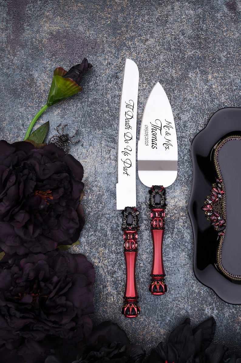 May include: A set of two silver cake knives with black and red jeweled handles. The knives are engraved with "Mr. & Mrs. Thomas 09/10/2023" and "Til Death Do Us Part".