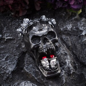 May include: A black skull-shaped decorative item with the text "A Till Death" and a floral crown. Two silver skull-shaped ring holders with rings featuring red and clear gemstones are inside the skull's mouth. The item is on a dark, textured surface.