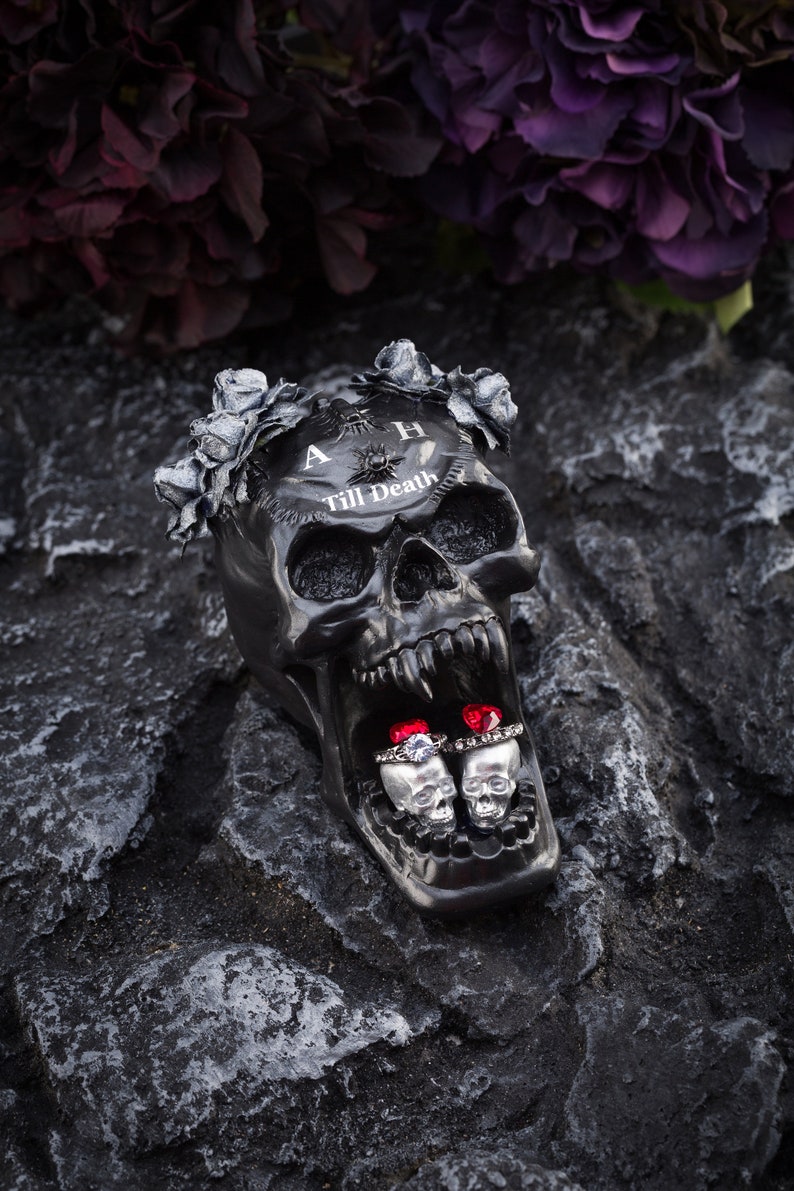 May include: A black skull-shaped decorative item with the text "A Till Death" and a floral crown. Two silver skull-shaped ring holders with rings featuring red and clear gemstones are inside the skull's mouth. The item is on a dark, textured surface.
