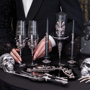 May include: Black and silver wedding cake topper set with two skulls, two champagne flutes, a cake knife, and a candle holder with a black candle. The flutes are decorated with silver skeletons and rhinestones. The candle holder is decorated with silver skeletons and rhinestones. The cake knife is black with a silver handle.