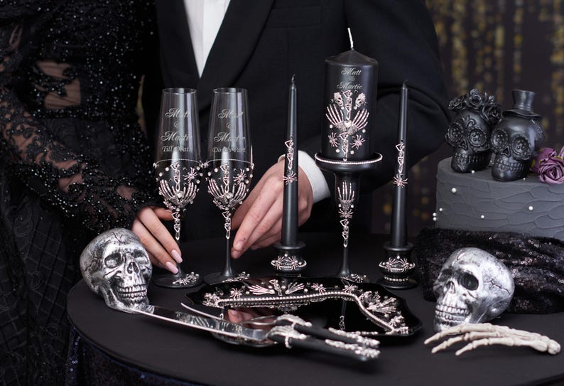 May include: Black and silver wedding cake topper set with two skulls, two champagne flutes, a cake knife, and a candle holder with a black candle. The flutes are decorated with silver skeletons and rhinestones. The candle holder is decorated with silver skeletons and rhinestones. The cake knife is black with a silver handle.