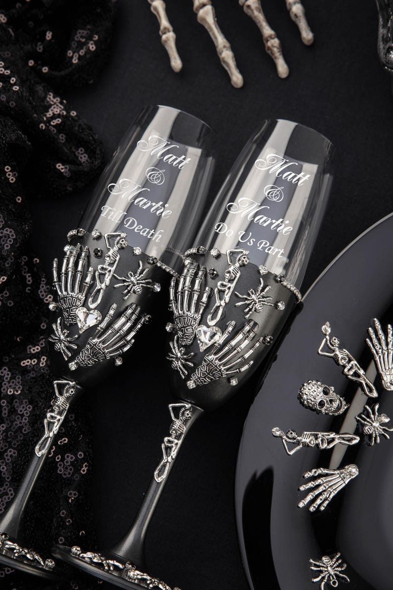May include: Two black champagne flutes with silver skeleton hands, spiders, and rhinestones. The flutes are engraved with the text "Matt & Martie Till Death" and "Matt & Martie Do Us Part".