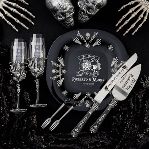 May include: A black wedding set with a skeleton theme. Includes two champagne flutes, a plate, cake server, and forks. The plate and server are engraved with names and a date. The flutes and server have skeleton hand details.