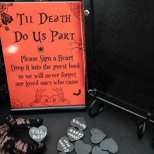 May include: A red sign with the text "Til Death Do Us Part" and "Please Sign a Heart" is displayed next to a black guest book. Small black heart-shaped tags with white lettering are scattered around. The theme is gothic wedding decor.