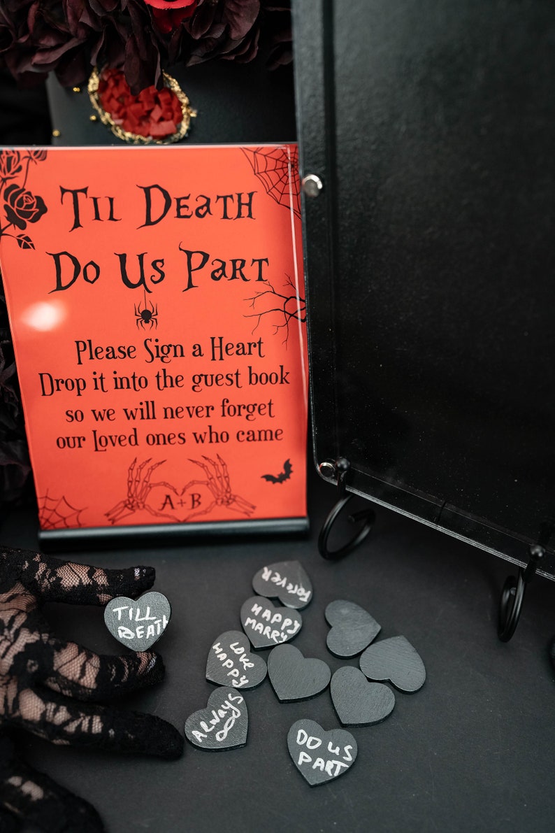 May include: A red sign with the text "Til Death Do Us Part" and "Please Sign a Heart" is displayed next to a black guest book. Small black heart-shaped tags with white lettering are scattered around. The theme is gothic wedding decor.