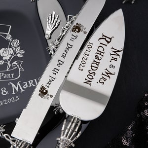 May include: A silver cake knife and server set with skeleton hand handles. The knife is engraved with "'Til Death Do Us Part" and a skull design. The server is engraved with "Mr. & Mrs. Richardson 10/13/2023". A black plate with a rose design is in the background.