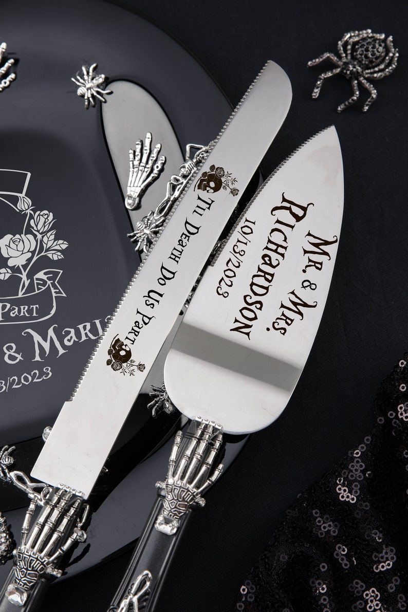 May include: A silver cake knife and server set with skeleton hand handles. The knife is engraved with "'Til Death Do Us Part" and a skull design. The server is engraved with "Mr. & Mrs. Richardson 10/13/2023". A black plate with a rose design is in the background.