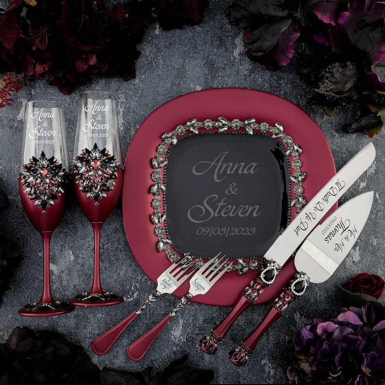 May include: A burgundy cake plate with a black square plate in the center. The black plate has the words "Anna & Steven" and the date "09/03/2023" engraved on it. Two burgundy champagne flutes with rhinestones and two burgundy cake knives with rhinestones are also included.