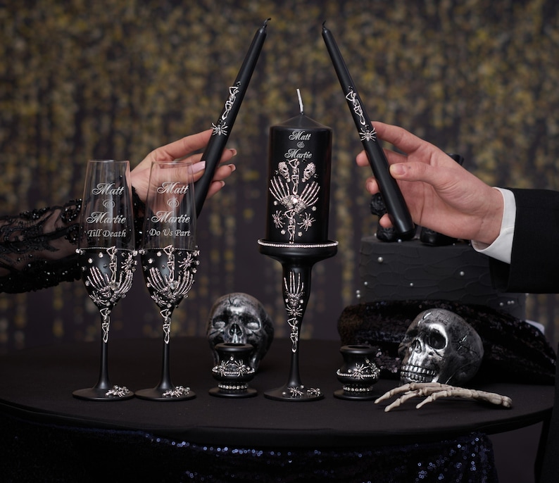 May include: Black wedding candle set with a skull and bone design. The candle is decorated with silver glitter and the words "Matt & Marie" are written on the side. Two black champagne flutes with silver glitter and the words "Till Death" and "Do Us Part" are also included.
