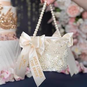 May include: Ivory wedding basket with pearl handle and floral beadwork. Features a satin bow with a pearl embellishment and a ribbon that reads "Here Comes The Bride" and "August 21, 2025".