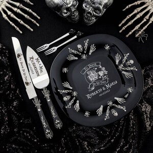 May include: A black wedding plate, cake server, and forks with a skeleton theme. The plate has the text "Til Death Do Us Part" and the names "Roberto & Maria" with the date 10/13/2023. The cake server and forks are silver.