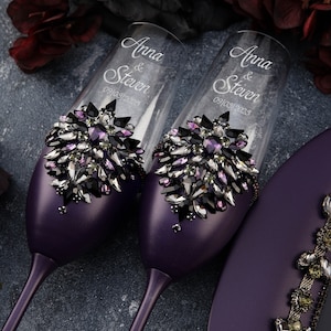 May include: Two purple champagne flutes decorated with black and purple rhinestones. The flutes are engraved with the names "Anna & Steven" and the date "09/03/2023".