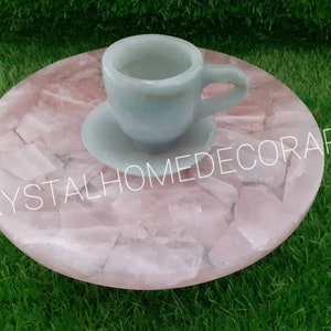 May include: A light blue ceramic teacup and saucer sitting on a pink rose quartz coaster. The coaster is made of many small pieces of rose quartz that have been glued together. The coaster is round and has a smooth surface. The teacup and saucer are both decorated with a simple design.