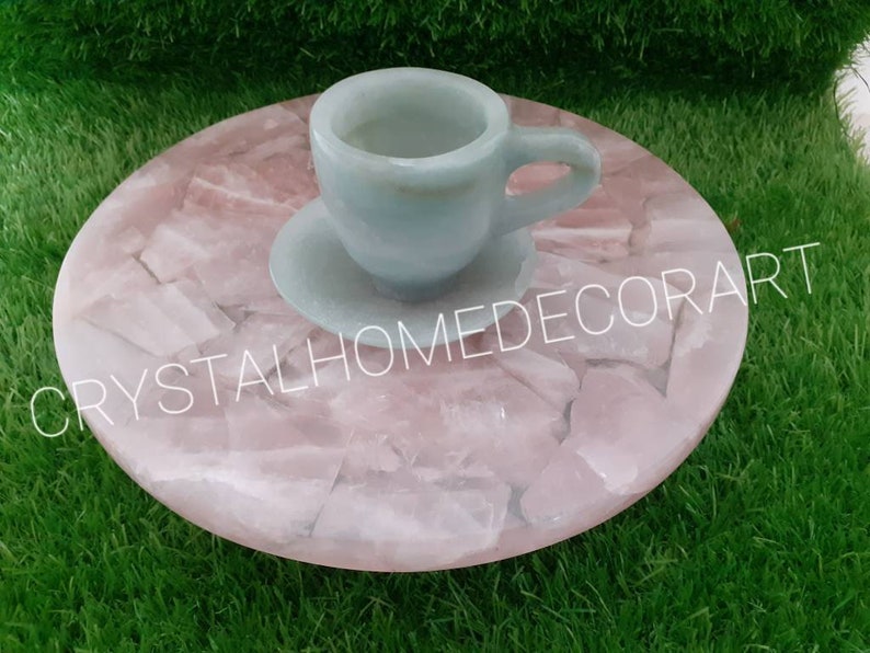 May include: A light blue ceramic teacup and saucer sitting on a pink rose quartz coaster. The coaster is made of many small pieces of rose quartz that have been glued together. The coaster is round and has a smooth surface. The teacup and saucer are both decorated with a simple design.