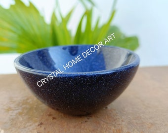 May include: A small, dark blue bowl made of polished lapis lazuli. The bowl has a speckled, glittery appearance.  The bowl is sitting on a brown surface.  "CRYSTAL HOME DECOR ART" is written in white text on the bowl.
