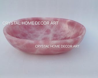 May include: A pink rose quartz bowl with a smooth, polished surface. The bowl has a rounded, oval shape and displays a natural, marbled pattern of pink and white. The text "CRYSTAL HOME DECOR ART" is printed on the image.