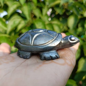 May include: A small, silver-coloured turtle figurine carved from hematite. The turtle is sitting on a person's hand.