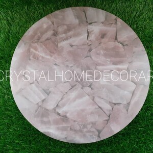May include: A round pink crystal table top made of many small pieces of rose quartz. The table top is on a green grass background.  CRYSTALHOMEDECORART is printed on the table top.