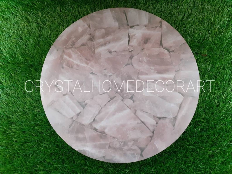 May include: A round pink crystal table top made of many small pieces of rose quartz. The table top is on a green grass background.  CRYSTALHOMEDECORART is printed on the table top.
