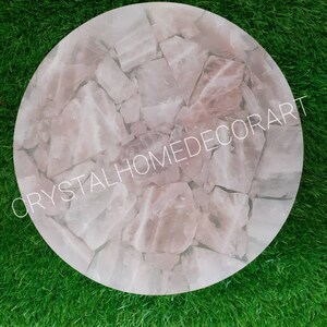 May include: A round, pink, crystal table top made of many small pieces of rose quartz. The table top is on a green grass surface. The text "CRYSTALHOMEDECORART" is visible in the image.
