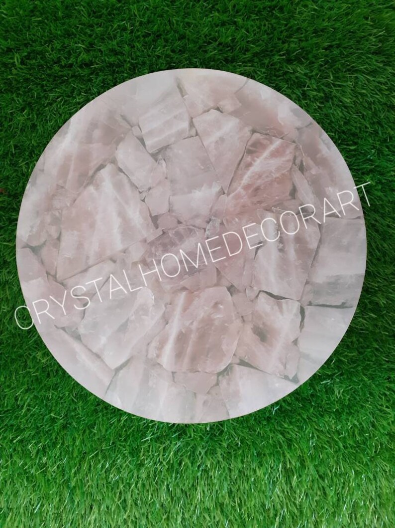 May include: A round, pink, crystal table top made of many small pieces of rose quartz. The table top is on a green grass surface. The text "CRYSTALHOMEDECORART" is visible in the image.
