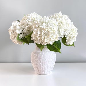 以下が含まれることがあります： 緑の葉がついた白いアジサイの花束が、質感のあるオフホワイトの陶器製花瓶に生けられています。花瓶は縦に溝が入った丸みを帯びた形状で、斑点模様の仕上げが施されており、大きな白い花びらの密集した房がいくつか活けられています。