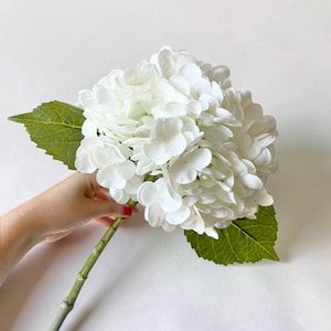 以下が含まれることがあります： 白い花びらが大きく丸く集まった人工のアジサイの茎。花頭は細い緑色の茎に取り付けられており、花の付け根からは2枚の目立つ質感のある緑色の葉が伸びています。