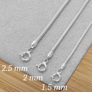 May include: Three silver snake chains of different widths, 1.5 mm, 2 mm, and 2.5 mm. The chains are laid out on a gray surface with a wooden surface visible in the background.