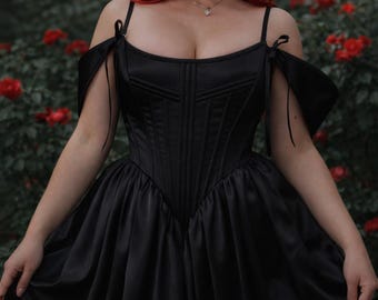 May include: A black satin corset dress with off-the-shoulder sleeves. The dress features a structured bodice and a flowing skirt. The model has red hair and is wearing a necklace. The background is a garden with red flowers.