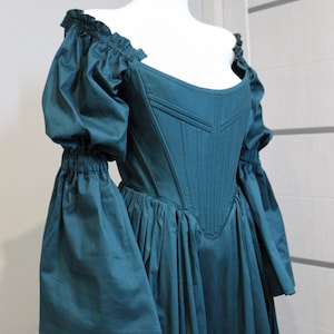 May include: Teal-colored historical dress with a fitted bodice and a flared skirt. The dress features off-the-shoulder, puffed sleeves gathered at the elbow. The bodice has vertical seams and a sweetheart neckline, displayed on a white mannequin.