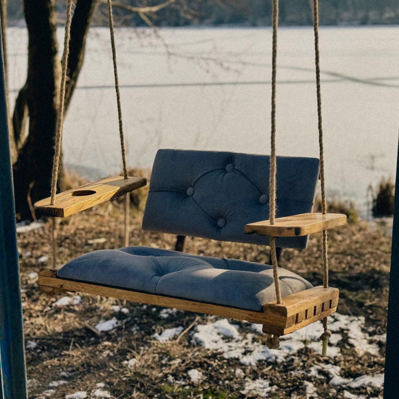 May include: A wooden swing seat with a blue velvet cushion and a wooden tray on each side. The swing is hanging from a tree branch and is in front of a lake.