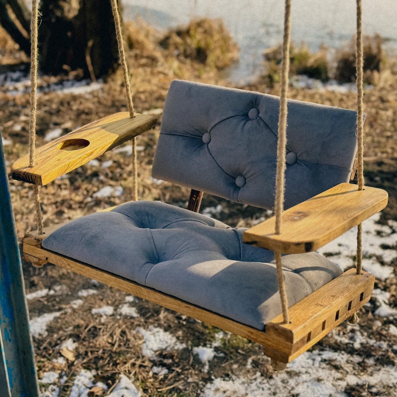 May include: A wooden swing with a gray cushioned seat and back. The swing has two wooden armrests and a small wooden shelf on the left side.