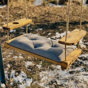 May include: A wooden swing with a gray cushioned seat and two wooden shelves. The swing is hanging from ropes and is outdoors.