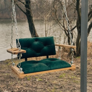May include: A wooden swing seat with a dark green velvet cushion and a small wooden side table. The swing is hanging from chains and is outdoors.