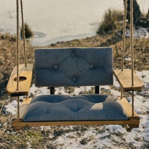May include: A gray cushioned swing seat with wooden arms and a wooden frame. The swing is hanging from two ropes.