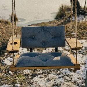 May include: A wooden swing seat with a gray cushioned seat and back. The swing has two side arms, one with a cup holder. The swing is hanging from two ropes.