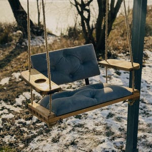 May include: A wooden swing chair with a gray fabric cushion and a built-in cup holder. The swing is hanging from a tree branch and is suspended by two ropes.