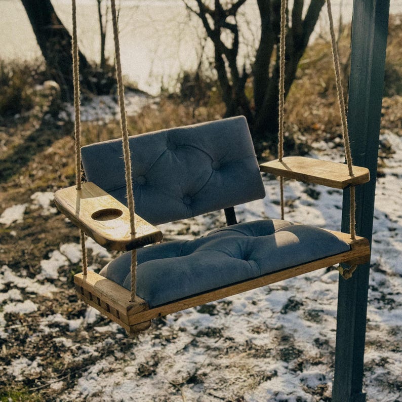 May include: A wooden swing chair with a gray fabric cushion and a built-in cup holder. The swing is hanging from a tree branch and is suspended by two ropes.